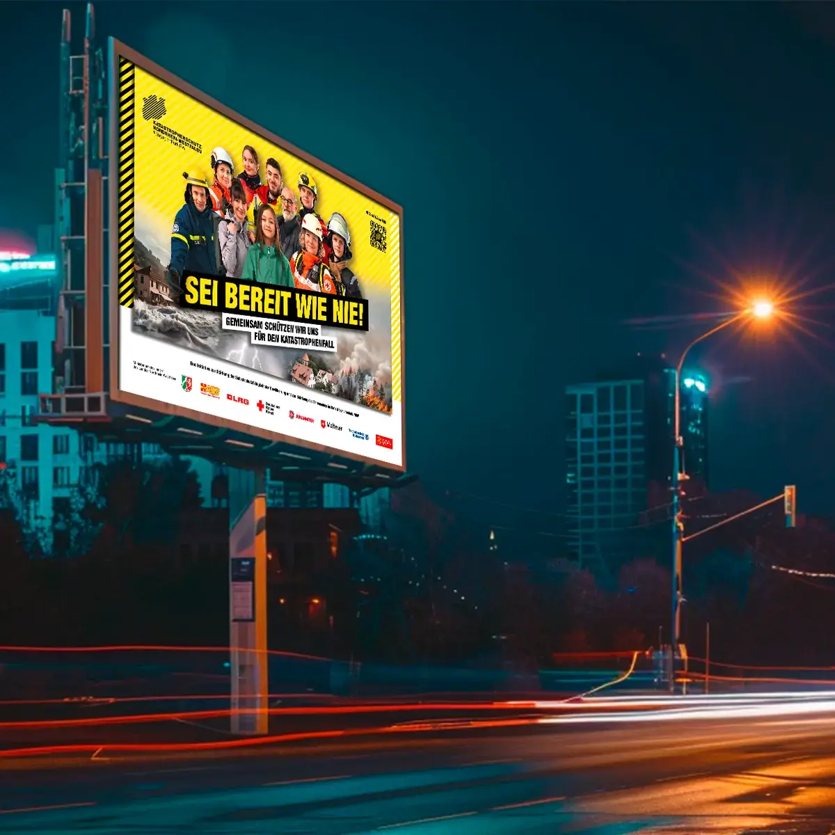 Das Bild zeigt eine große Plakatwand an einer Straße. Auf dem Plakat: Eine Gruppe aus Katastrophenschützerinnen und Zivillisten stehen vor einem gelben Hintergrund. Auf dem Bild steht der Slogan &quot;SEI BEREIT WIE NIE!&quot;