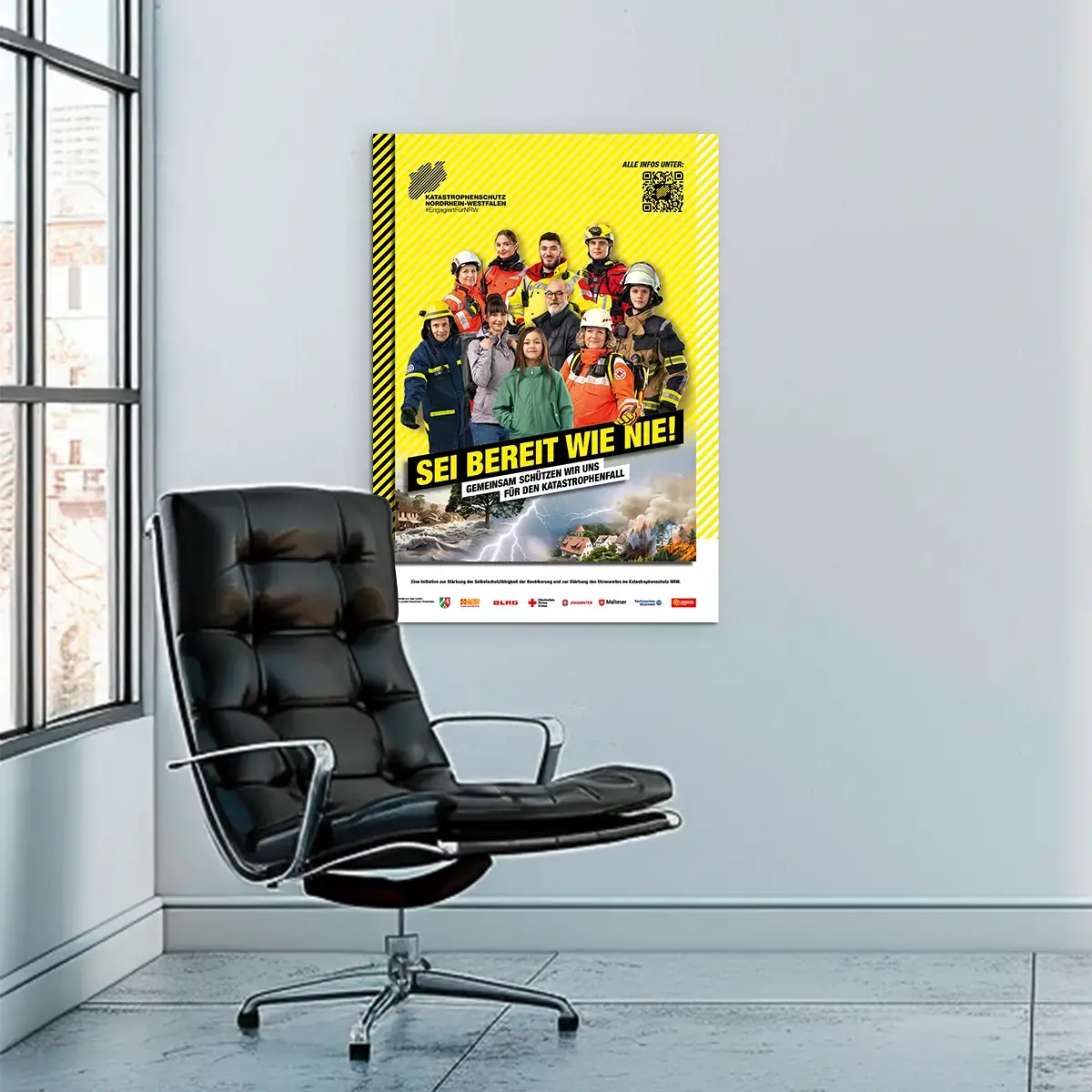 Das Bild zeigt ein Büro, in dem ein Stuhl vor einem Poster abgebildet ist. Auf dem Bild:Eine Gruppe aus Katastrophenschützerinnen und Zivillisten stehen vor einem gelben Hintergrund. Auf dem Bild steht der Slogan &quot;SEI BEREIT WIE NIE!&quot;