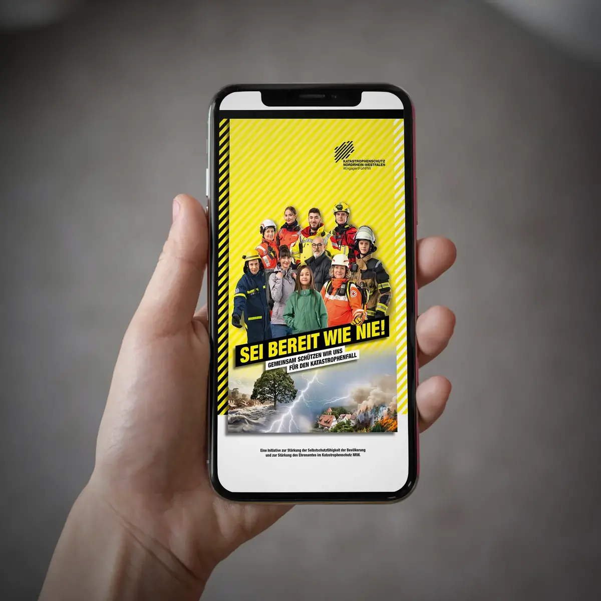Das Bild zeigt ein Smartphone, das ein Bild von einem Social-Media-Post darstellt. Auf dem Bild:Eine Gruppe aus Katastrophenschützerinnen und Zivillisten stehen vor einem gelben Hintergrund. Auf dem Bild steht der Slogan &quot;SEI BEREIT WIE NIE!&quot;